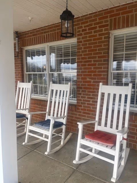 Three white wooden rocking chairs with cushions on a covered porch in front of a brick wall with two windows. A black hanging lantern light fixture is above the chairs.