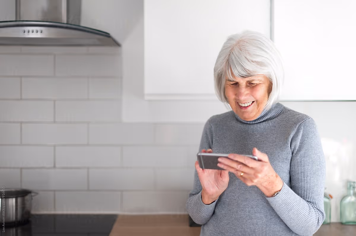 An elderly woman with white hair wearing a gray sweater is standing in a modern kitchen, smiling and looking at a smartphone she is holding in her hands. The kitchen has white tiled walls, a stainless steel range hood, and a stovetop with a pot on it.