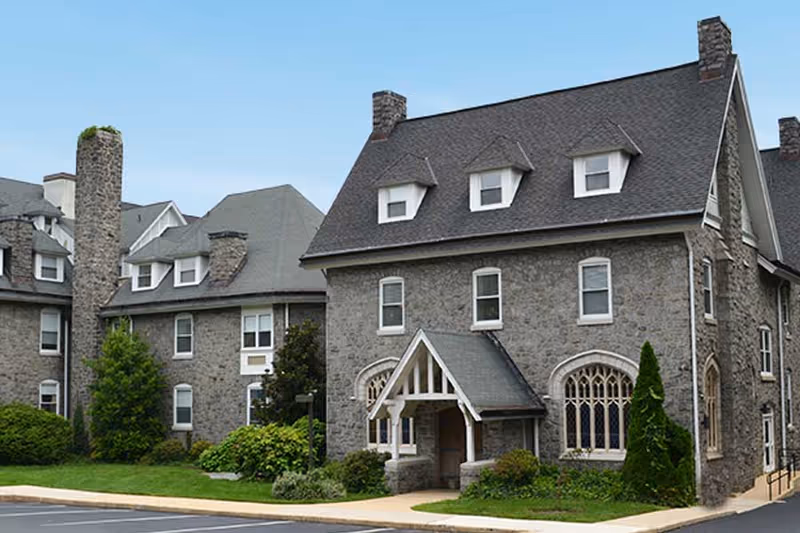 Exterior view of a large stone building with multiple chimneys, dormer windows, and a covered entrance, surrounded by greenery and a paved driveway under a clear blue sky.
