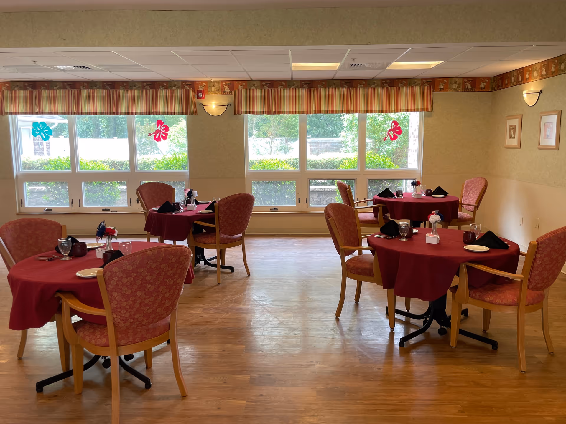 A dining room with four round tables covered with red tablecloths, each set with plates, glasses, napkins, and small flower centerpieces. The room has large windows with striped valances, letting in natural light and showing greenery outside. The walls are light-colored with framed pictures and wall sconces providing additional lighting. The floor is wooden.