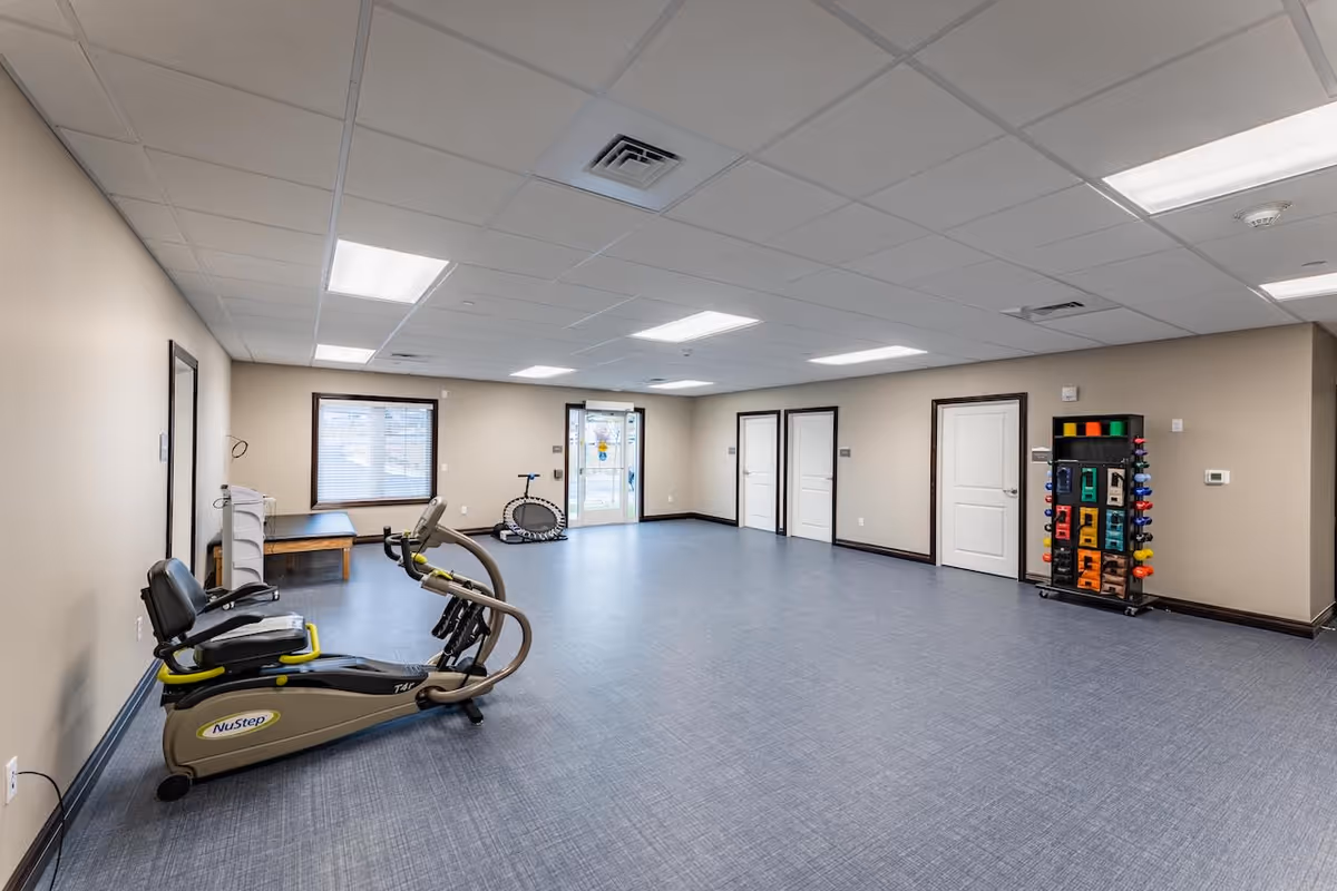 A spacious exercise room with light beige walls and a blue-gray floor. The room contains a NuStep recumbent cross trainer, a small trampoline, a bench, and a rack with colorful hand weights. There are several closed white doors along one wall and a window with blinds on another. The ceiling has recessed lighting panels.