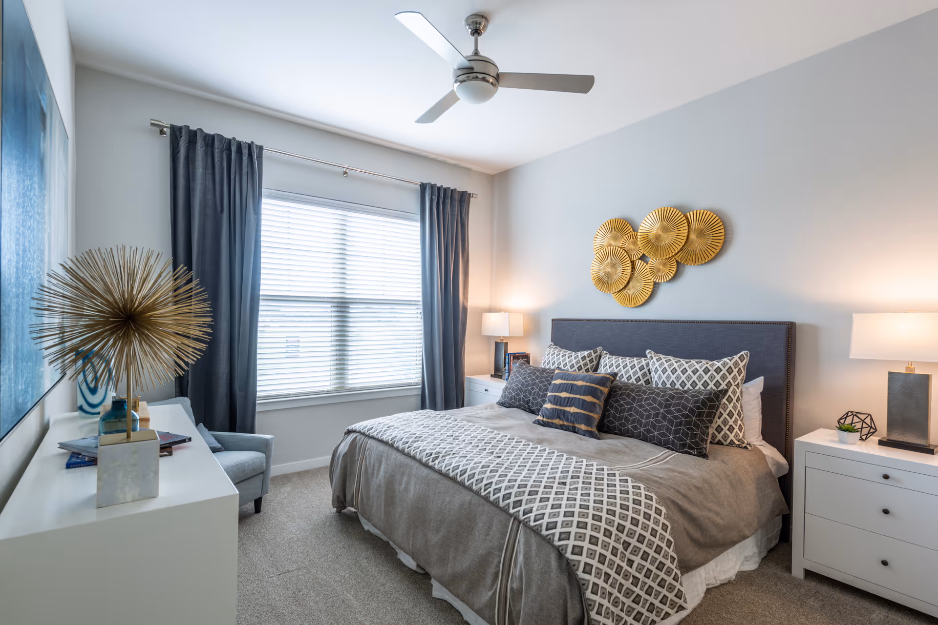 A neatly made bed with multiple decorative pillows and a patterned throw blanket in a bedroom. The room has light gray walls, a ceiling fan, two white nightstands with lamps, a window with gray curtains, and a white dresser with decorative items including a gold spiky sculpture.