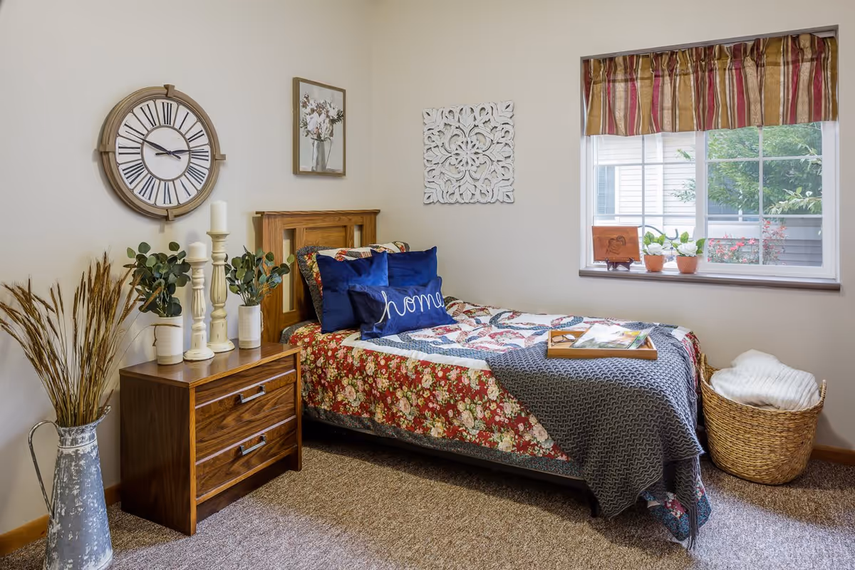 A cozy bedroom with a single bed covered in a floral quilt and blue pillows, one of which has the word 'home' embroidered on it. Next to the bed is a wooden nightstand with three white candles and two small potted plants. A large round wall clock and framed artwork hang on the wall above the nightstand. A window with striped curtains lets in natural light, and a woven basket with a folded blanket sits on the floor near the window.