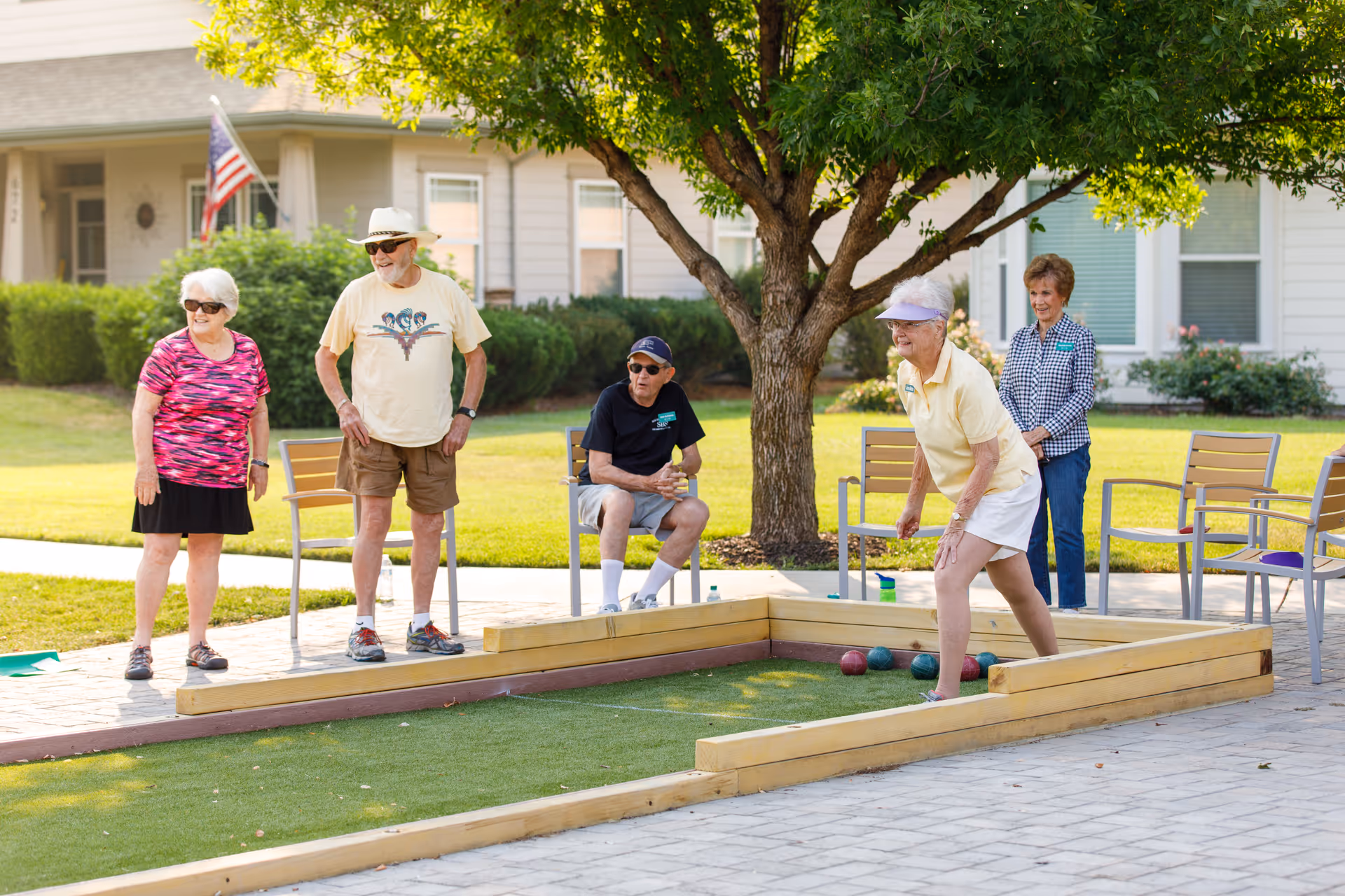 A group of five elderly people enjoying a game of bocce ball outdoors on a sunny day. They are standing and sitting around a bocce ball court with a tree providing shade. The background shows a residential building with windows and an American flag.
