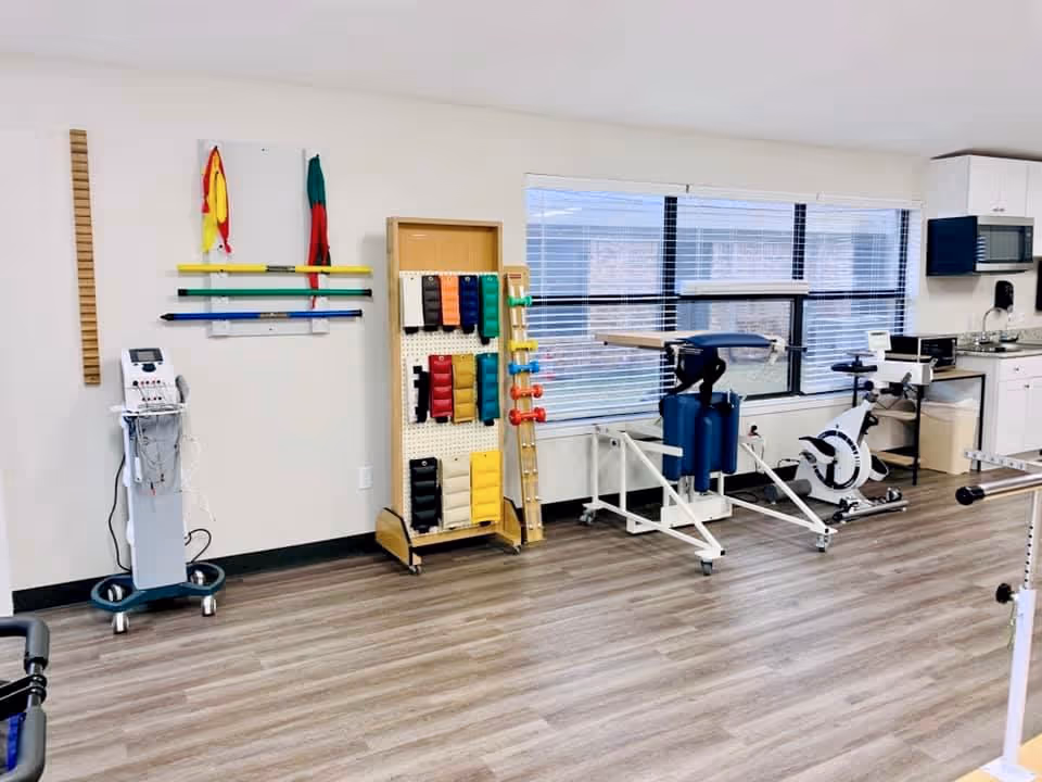 Bright rehabilitation/therapy room with exercise equipment, therapy table, free weights and a kitchenette along a windowed wall.