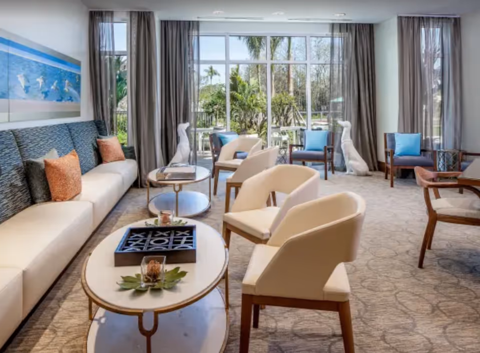 A bright and modern living room with large windows showing a garden outside. The room features a long white sofa with colorful pillows, several white and wooden chairs, two oval coffee tables with decorative items including a tic-tac-toe game, and sheer curtains framing the windows.