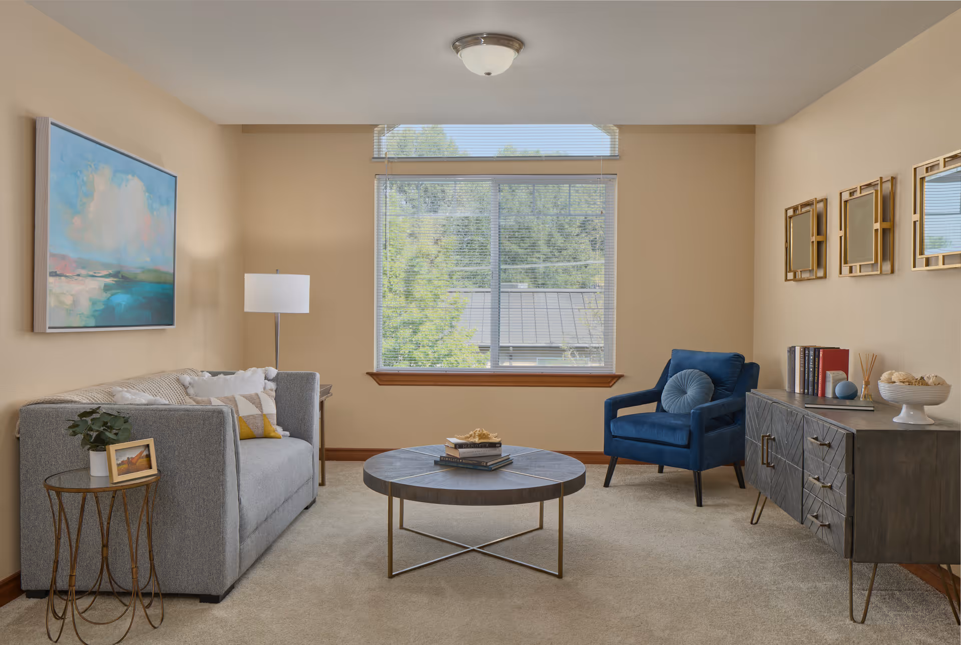 A cozy living room with a large window showing greenery outside. The room features a gray sofa with pillows, a blue armchair with a round cushion, a round coffee table with books and decorative items, a side table with a plant and framed photo, a floor lamp, and a dark wooden sideboard with books and decor. Three gold-framed mirrors hang on the wall above the sideboard.