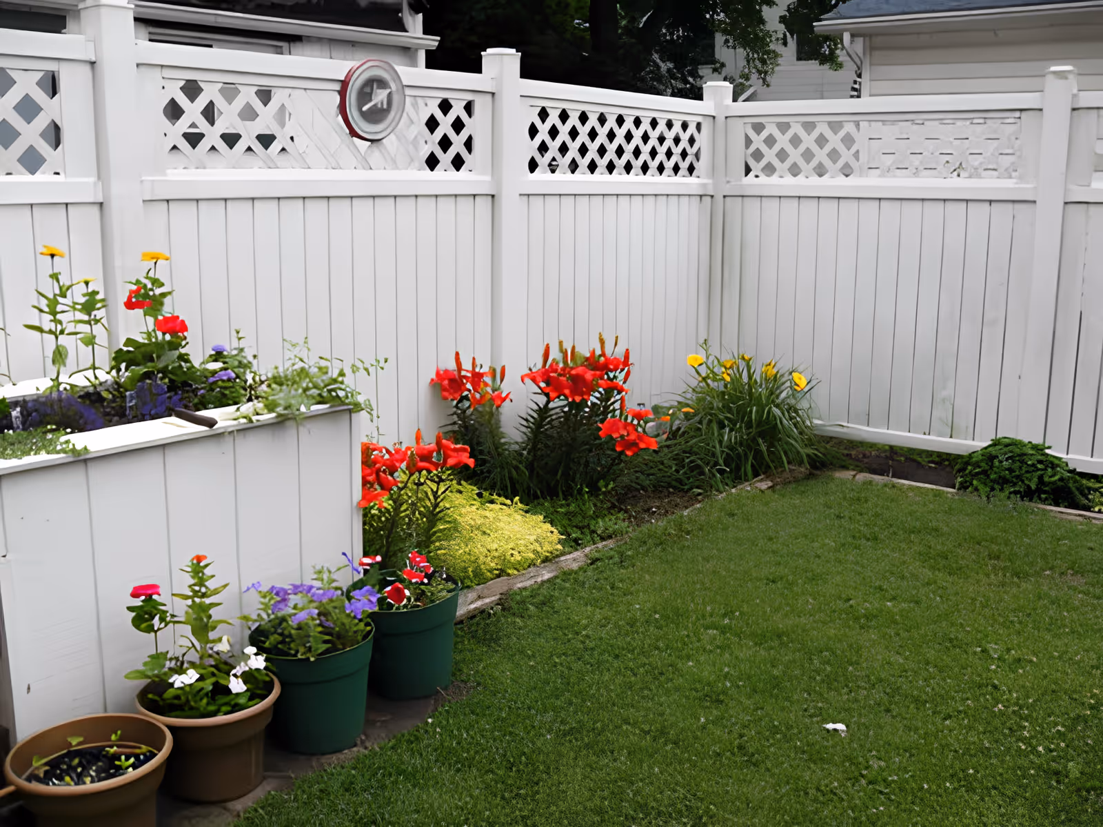A small garden area enclosed by a white wooden fence with lattice panels at the top. The garden features a variety of colorful flowers including red, yellow, purple, and white blooms planted in both the ground and pots. The grass lawn is well-maintained and green.