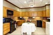 A kitchen area with wooden cabinets, a black microwave, black stove, black dishwasher, and a white washing machine. There is a round table in the center covered with a white tablecloth and a vase with flowers on top. The walls have a beige tile backsplash and the ceiling has recessed lighting.