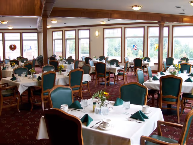 A bright dining room with multiple round tables covered in white tablecloths, each set with green napkins, cups, glasses, and small flower arrangements. The room has large windows letting in natural light and green cushioned wooden chairs around the tables.
