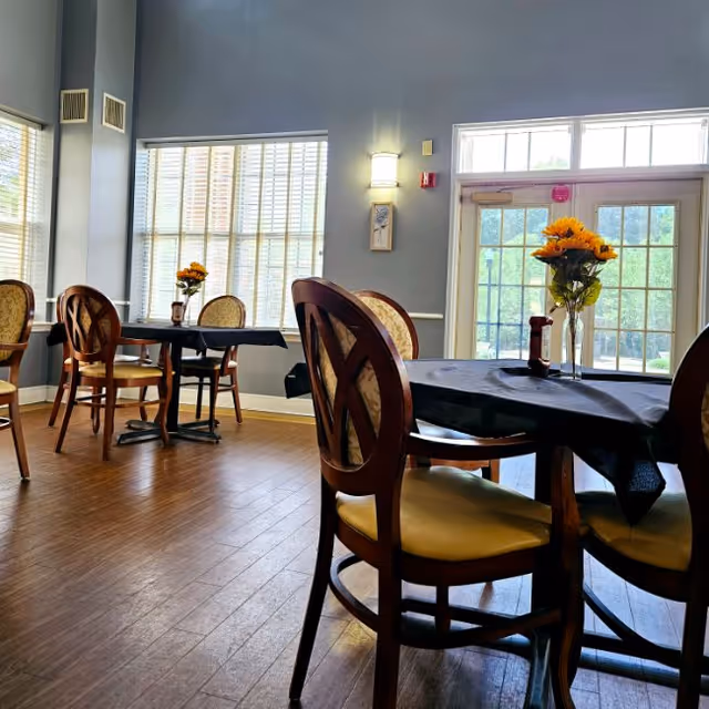 Dining room with round tables draped in black tablecloths, wooden chairs, vases of sunflowers, and large windows letting in natural light.