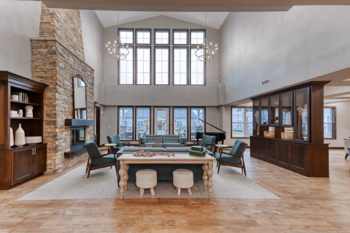 Spacious senior living facility common area with high ceilings and large windows. The room features a stone fireplace with a mirror above it, a wooden bookshelf with decorative items, a seating area with teal armchairs and sofas around a wooden coffee table, and a grand piano near the windows. The floor is wooden with a large area rug under the seating area, and two chandeliers hang from the ceiling.