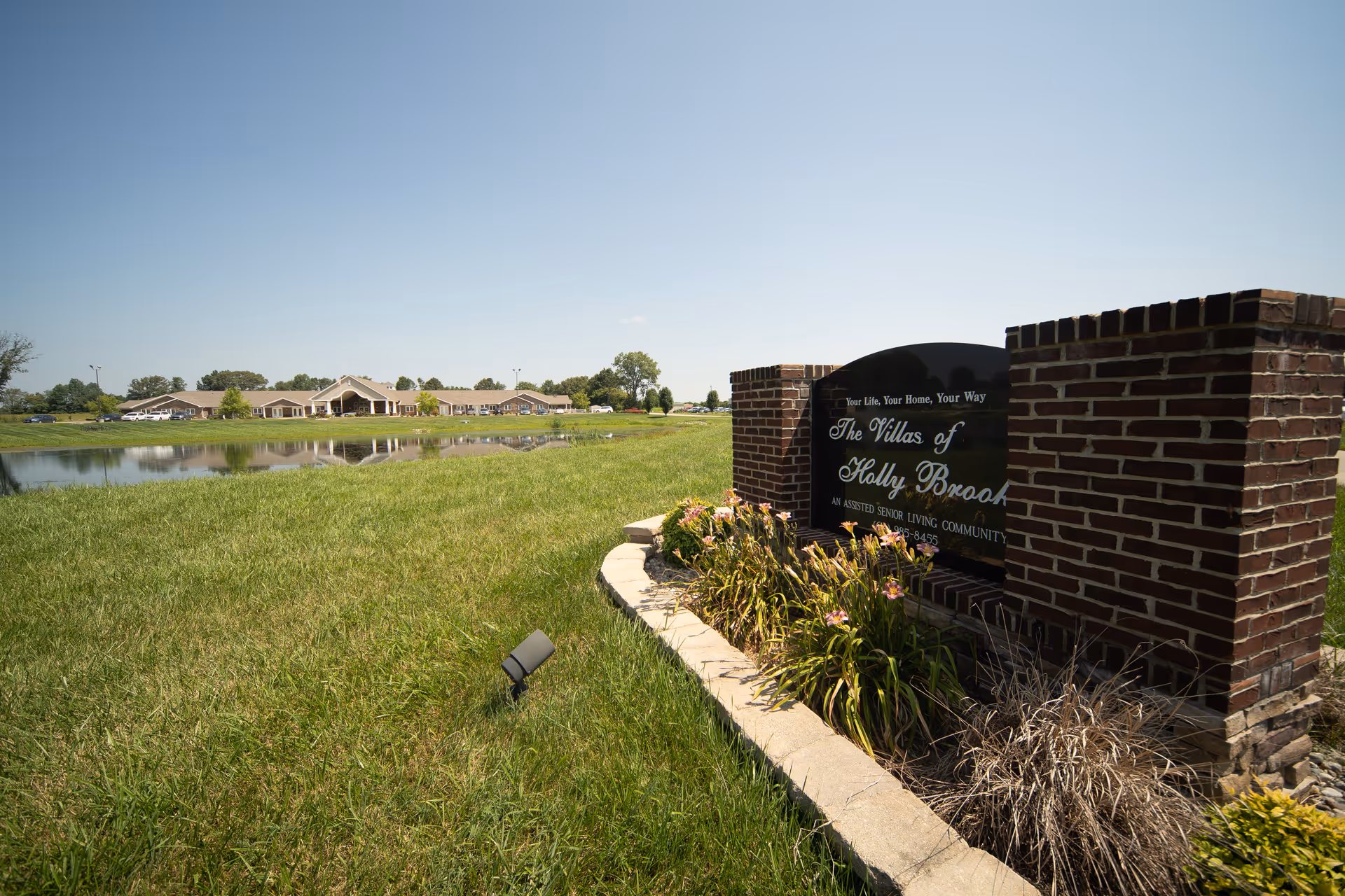 A grassy outdoor area with a pond reflecting a large single-story building in the background. In the foreground, there is a brick sign with flowers around it that reads 'The Villas of Holly Brook, An Assisted Senior Living Community.' The sky is clear and blue.