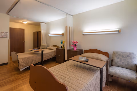 A shared bedroom in a senior living facility with two single beds, each with a beige quilted bedspread and an overbed table. There is a small dresser between the beds with a vase of pink flowers and a wall-mounted light above each bed. A patterned armchair is placed next to one of the beds. The room has wood flooring, a closed door, and a ceiling-mounted privacy curtain rail.