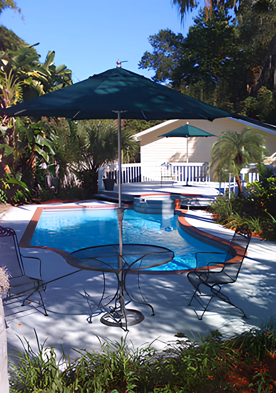 Outdoor swimming pool area with a round glass table and metal chairs under a large green umbrella. The pool is surrounded by lush greenery and plants, with a white deck and a small building in the background also featuring a green umbrella.