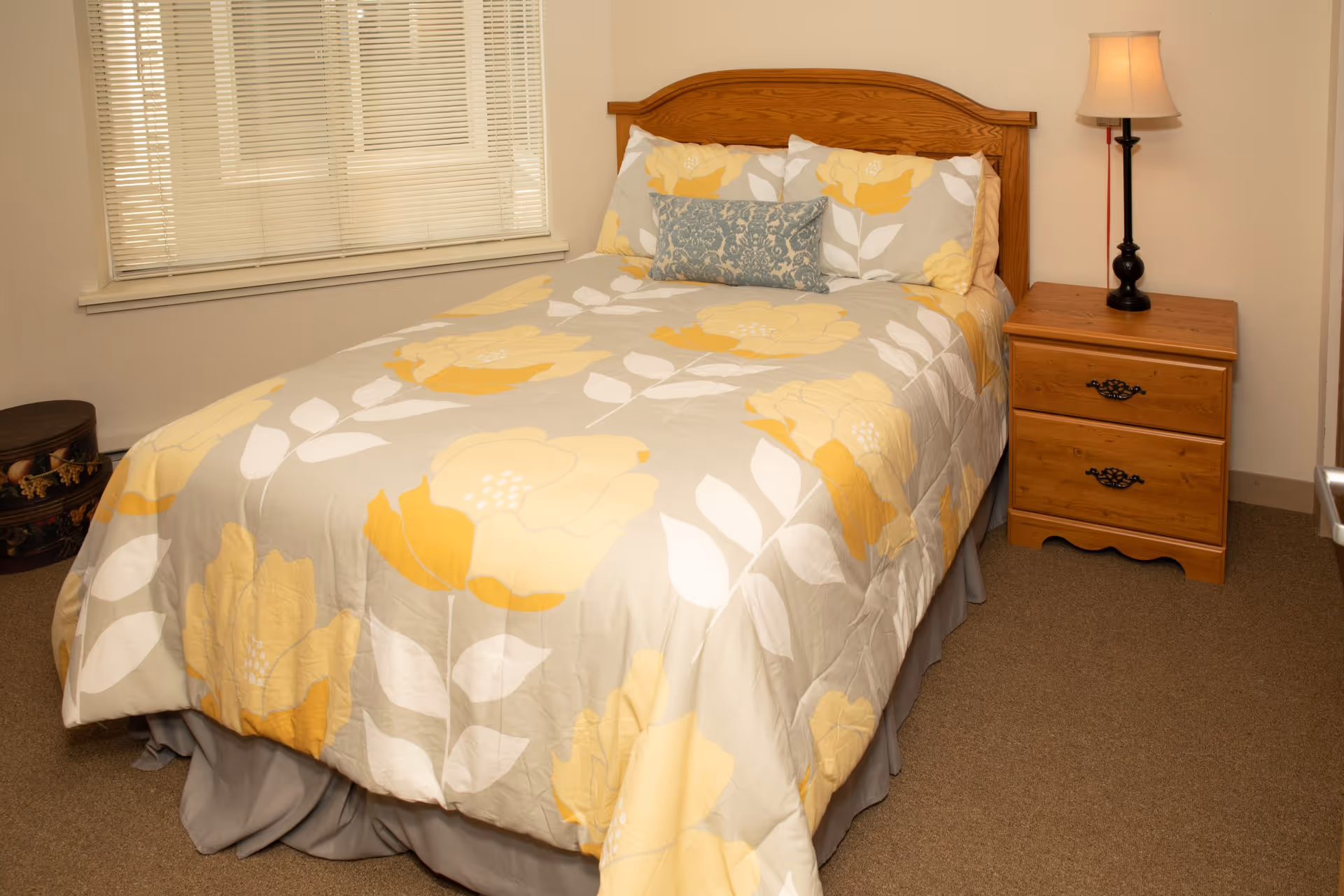 A bedroom with a wooden bed frame and a bed made with a floral-patterned comforter in shades of yellow, white, and gray. There is a matching pillow and a decorative blue pillow on the bed. Next to the bed is a wooden nightstand with two drawers and a black lamp with a beige lampshade. A window with closed blinds is visible behind the bed, and a small round decorative item is placed on the floor near the window.