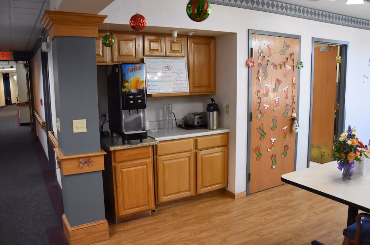 A small kitchenette area in an assisted living facility with wooden cabinets, a juice dispenser, a toaster, and a coffee pot on the countertop. Above the counter is a whiteboard with a menu written on it. To the right, a door decorated with Christmas stockings and candy canes is visible. A table with a vase of flowers is partially visible in the foreground, and a hallway extends to the left with an exit sign at the end.