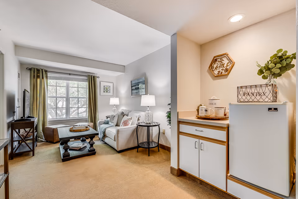 Bright furnished living room with a sofa, coffee table, TV and a small kitchenette area with a mini fridge and pantry decor.