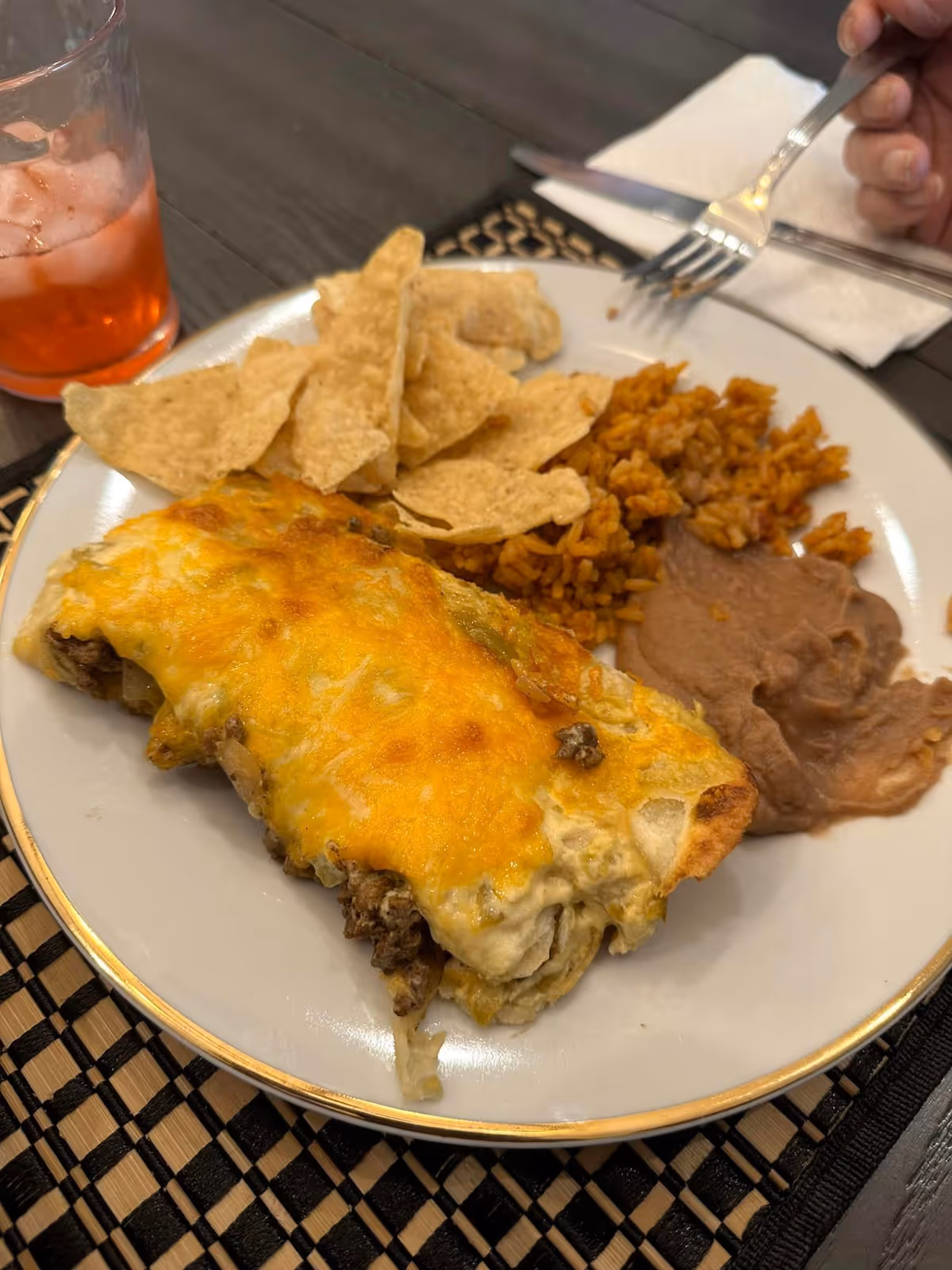 A plate of food featuring a cheesy baked dish, tortilla chips, Mexican rice, and refried beans, with a glass of iced beverage and a hand holding a fork in the background.