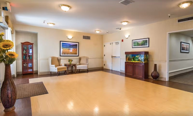 A bright senior living facility seating area with two armchairs, a small table, an aquarium, framed artwork, and decorative vases.