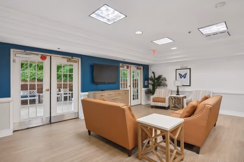 A bright and modern common area in a senior living facility featuring two tan leather sofas facing a wall-mounted flat screen TV above a wooden cabinet. There are two striped armchairs with a small table and lamp between them, a framed butterfly picture on the wall, and a potted plant in the corner. Double glass doors lead to an outdoor patio with seating visible through the doors. The room has wood flooring, white walls with blue accent panels, and recessed ceiling lights.