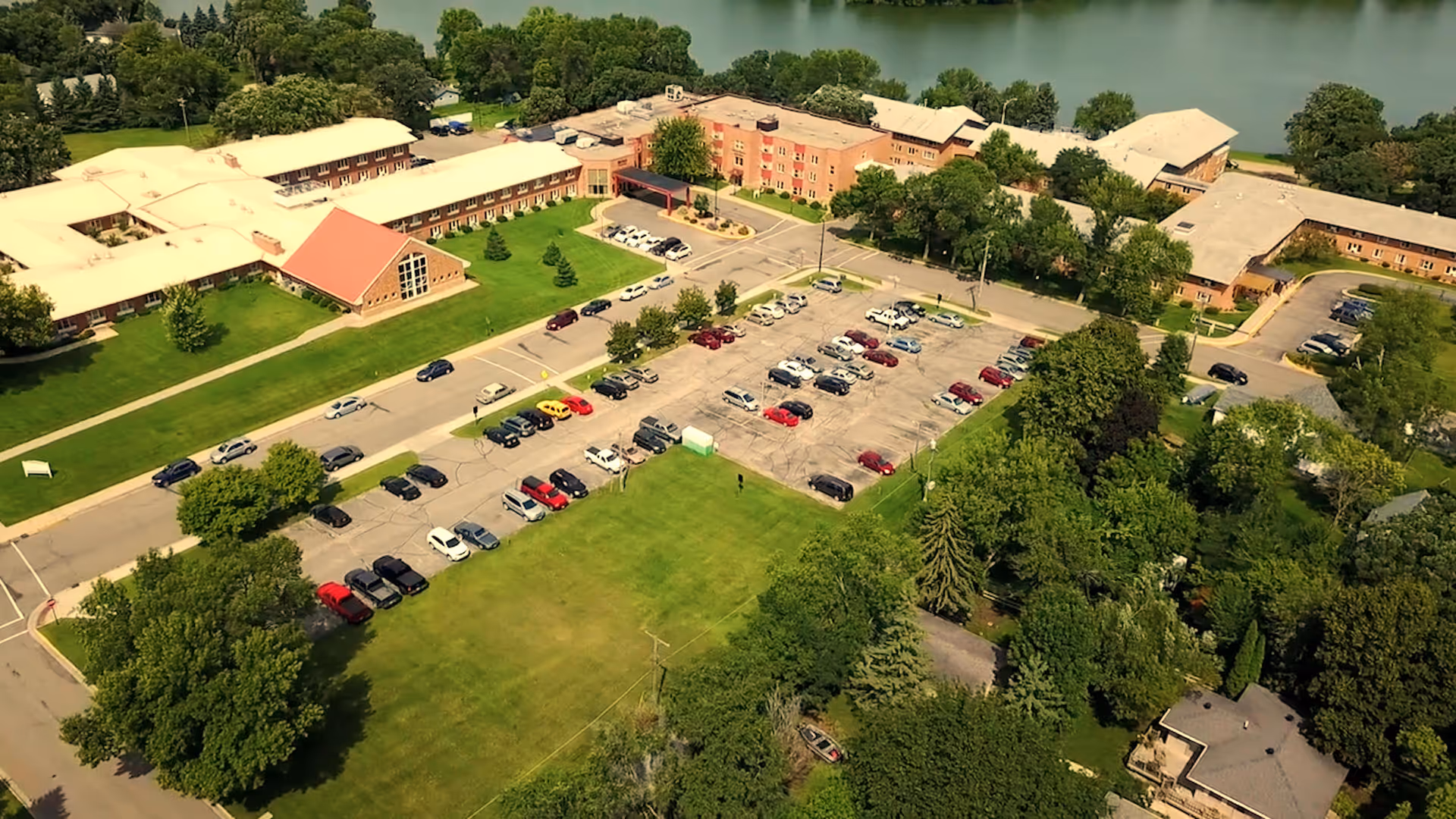 Aerial view of Bethany on the Lake senior living facility showing multiple connected buildings with beige roofs, a large parking lot filled with cars, green lawns, numerous trees, and a lake in the background.