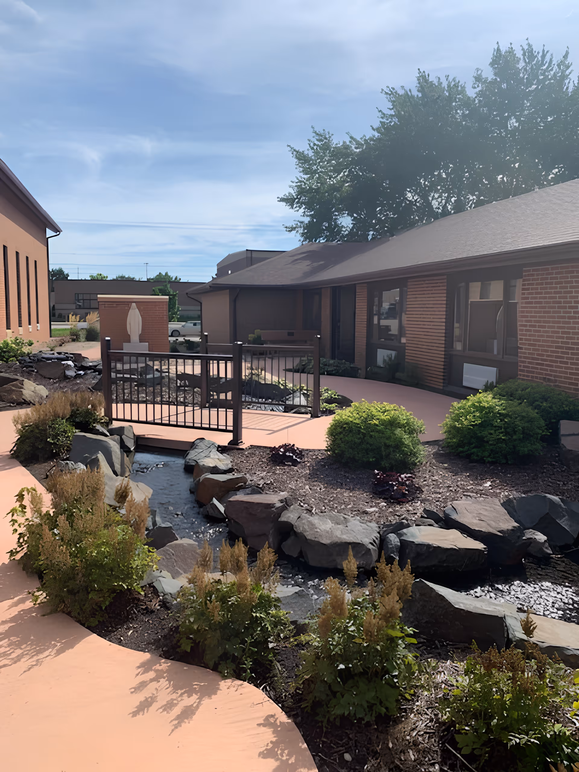 Outdoor garden area at Marian Home and Village featuring a small stream bordered by rocks and plants, a curved walkway, a black metal railing bridge, and brick buildings in the background under a partly cloudy sky.