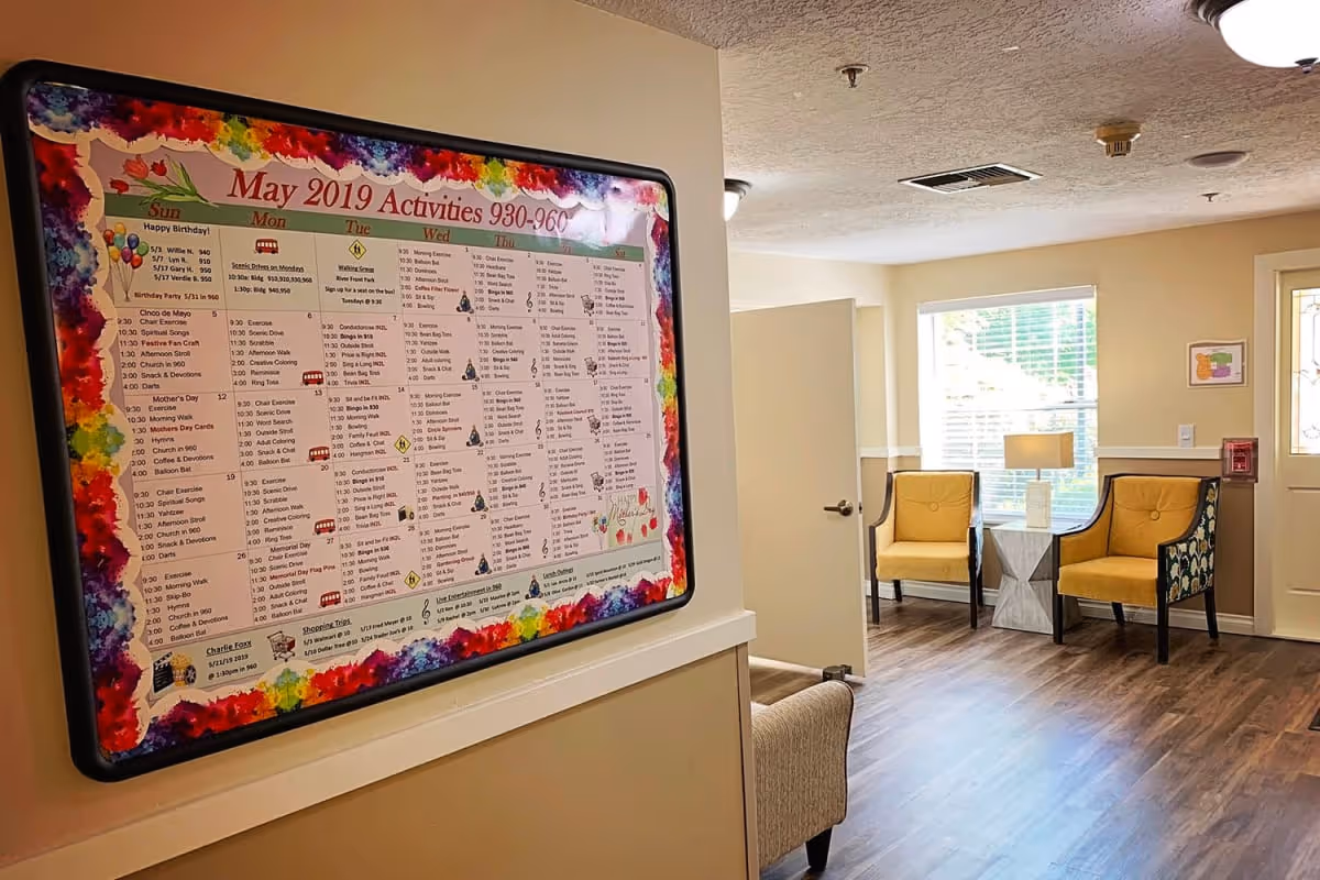 Interior view of a senior living facility hallway with a colorful May 2019 activities calendar on the wall. The hallway leads to a small sitting area with two yellow armchairs and a small table with a lamp, next to a window and a door.