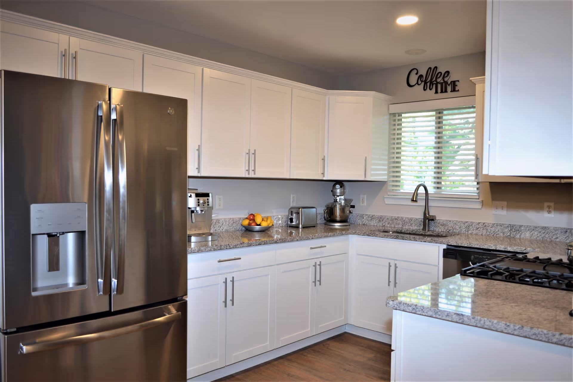 Modern kitchen with white cabinets, granite countertops, stainless steel refrigerator, stove, and dishwasher. A window with blinds is above the sink, and a decorative sign reading 'Coffee Time' is mounted on the wall above the window. Various kitchen appliances and a bowl of fruit are on the countertops.