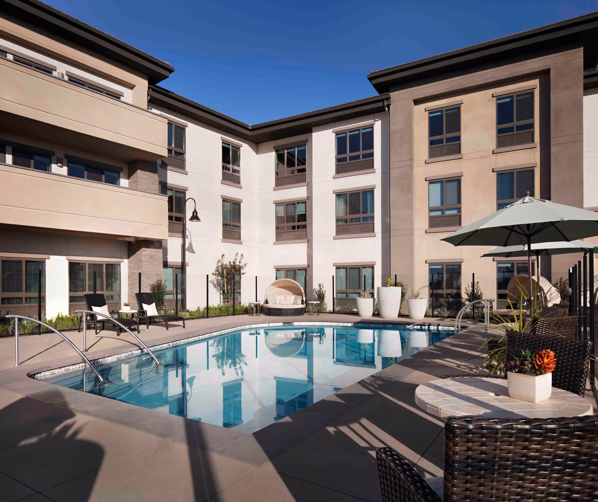 Outdoor swimming pool area at Belmont Village Senior Living Aliso Viejo with lounge chairs, tables with umbrellas, potted plants, and a three-story building in the background under a clear blue sky.
