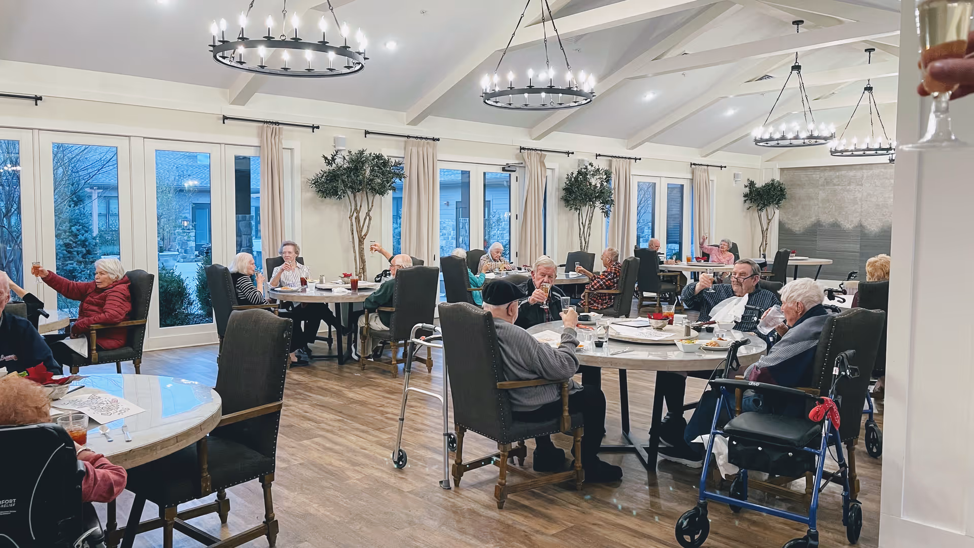 A spacious dining room in an assisted living facility with multiple round tables where elderly residents are seated, enjoying drinks and socializing. The room has large windows with beige curtains, wooden flooring, and modern circular chandeliers hanging from a white ceiling with exposed beams. Some residents use walkers and wheelchairs.