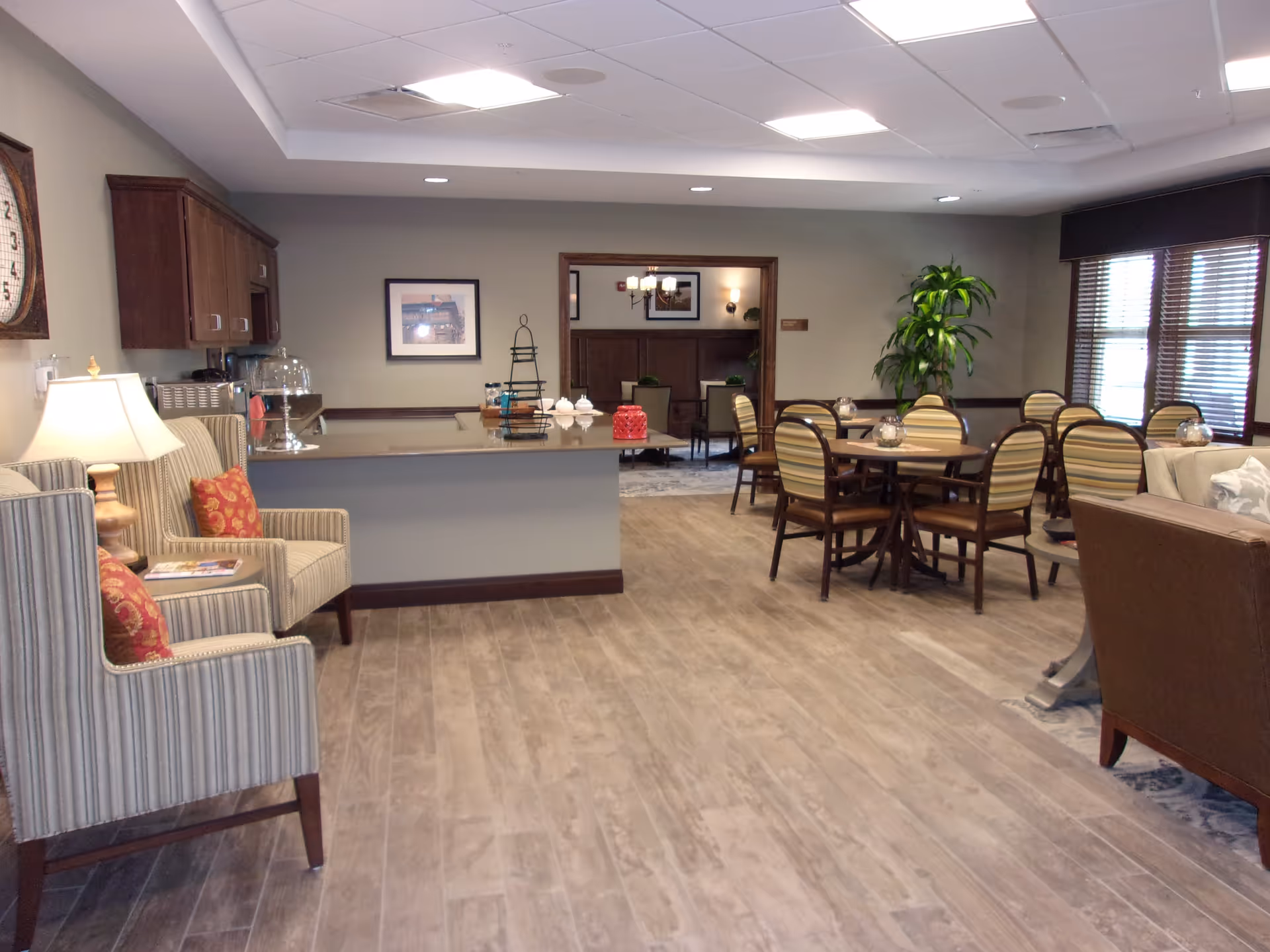 A spacious, well-lit common area in a senior living facility featuring a seating area with striped armchairs and red cushions, a countertop with cabinets above, round tables with striped chairs, a large green plant near the window with blinds, and a glimpse into an adjoining room with additional seating and framed pictures on the walls.