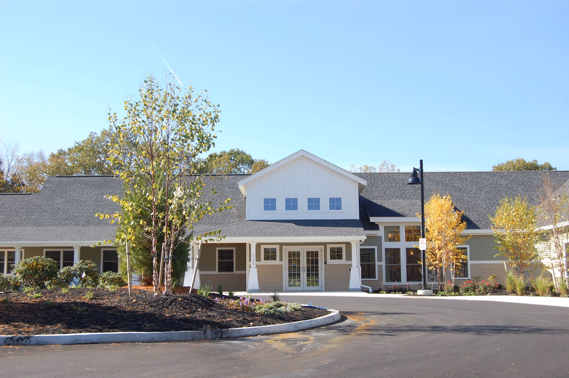 Front exterior view of a single-story building with a peaked roof, large windows, and a covered entrance. There are landscaped areas with trees and shrubs in front of the building, and a clear blue sky overhead.