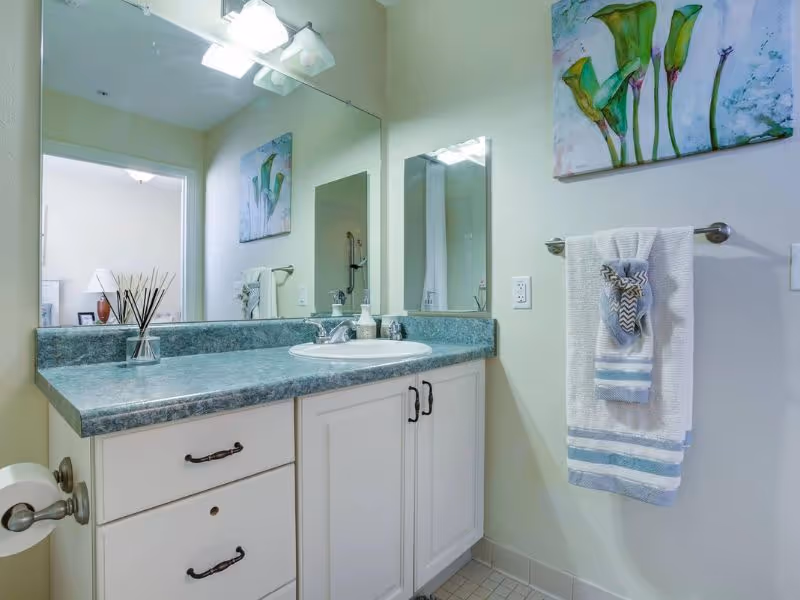 A bathroom vanity with a greenish countertop, a white sink, and white cabinets with dark handles. Above the sink is a large mirror reflecting part of the room, including a doorway and a lamp. On the wall to the right, there is a towel rack with neatly folded white and blue towels and a decorative painting of green flowers above it. The bathroom has light-colored walls and tiled flooring.