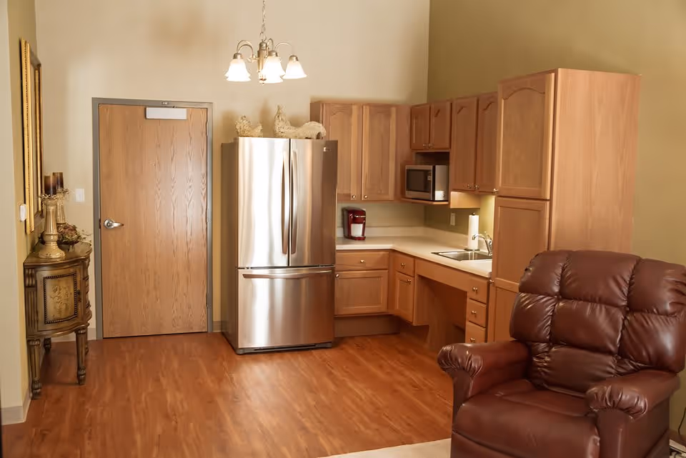 Small open kitchen area with a stainless steel refrigerator, wooden cabinets, sink, and a brown leather recliner in the foreground.