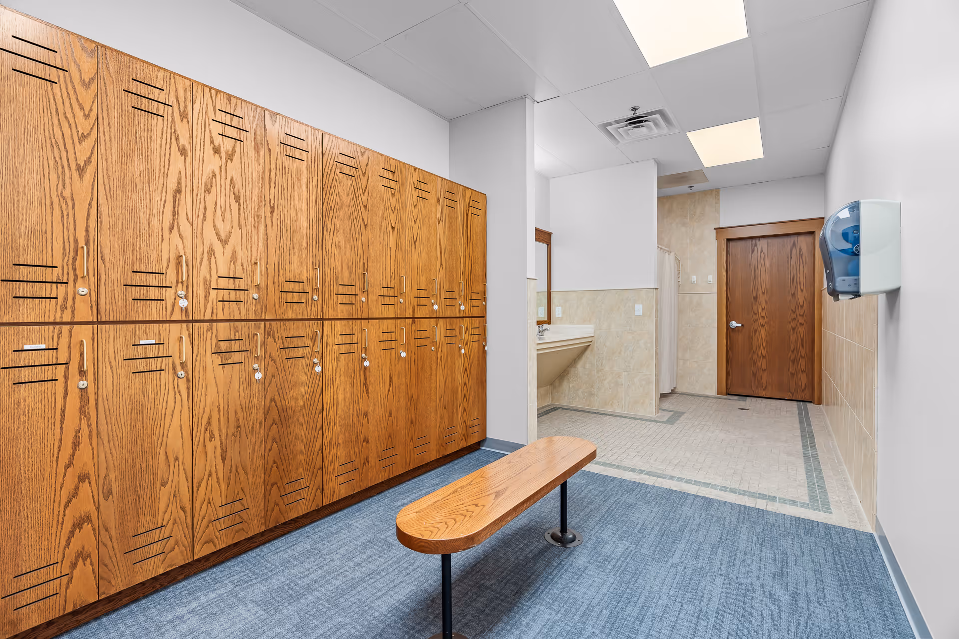 Interior view of a locker room with wooden lockers on the left side and a wooden bench in the center. The floor is carpeted in blue near the lockers and tiled near the sink area. There is a sink with a mirror on the far wall and a wooden door next to it. A paper towel dispenser is mounted on the right wall.