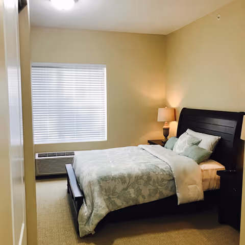 A neatly made bed with floral patterned bedding and multiple pillows in a bedroom with beige walls and carpet. There is a window with closed blinds and an air conditioning unit below it. A nightstand with a lamp is positioned next to the bed.