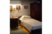 A small residential healthcare bedroom with a single bed, nightstand, lamp, framed pictures, and a wardrobe.
