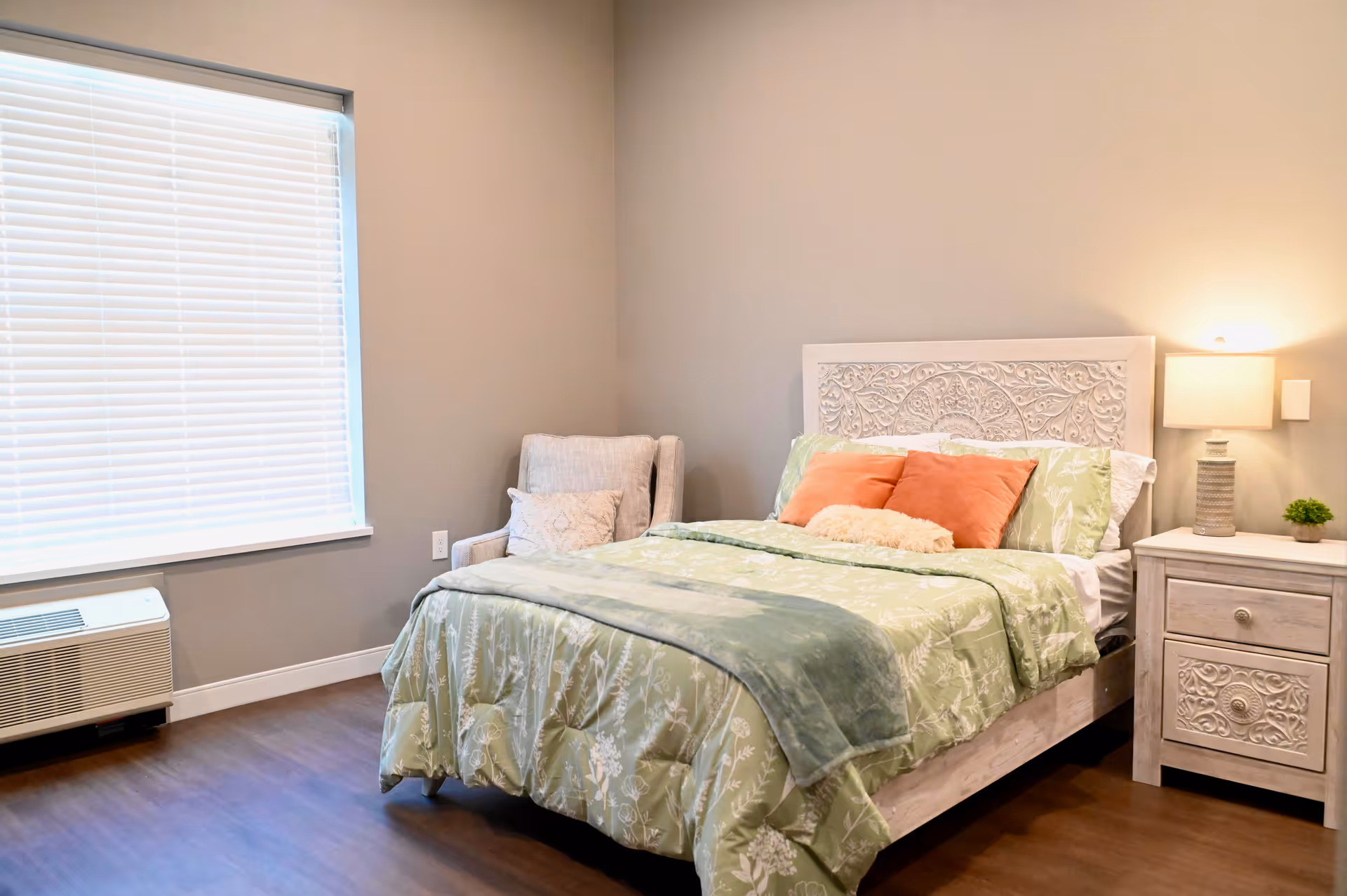 A tidy bedroom featuring a carved white headboard, green floral bedding, a nightstand with a lamp, and an upholstered chair by a window.