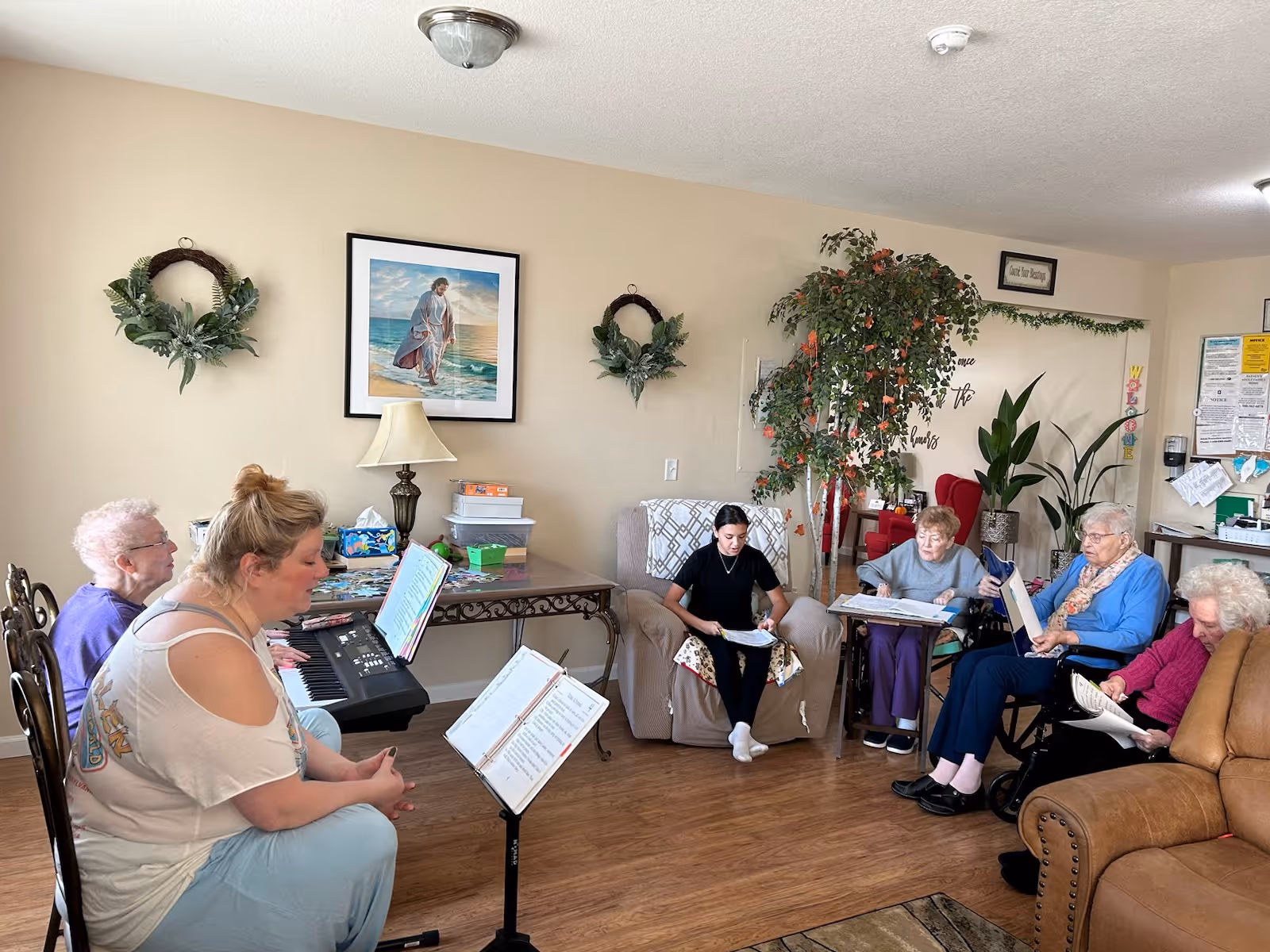 A group of elderly women and a younger woman gathered in a living room setting. One woman is playing a keyboard while others are seated on chairs and a couch, holding papers or books, possibly engaged in a group activity or music session. The room has beige walls decorated with wreaths, a framed picture, a lamp on a table, and several plants.