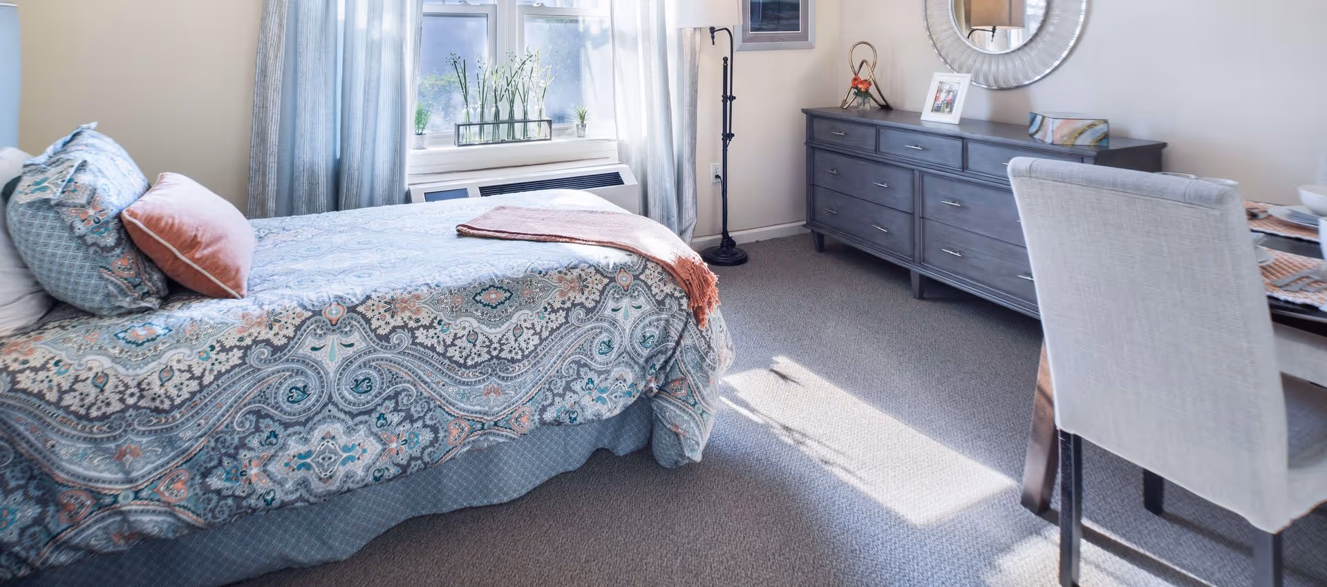 A bright bedroom with a bed covered in a patterned blue and coral bedspread, multiple pillows, a window with light blue curtains, a dark wooden dresser with a round mirror above it, a floor lamp, and a dining table with upholstered chairs partially visible.