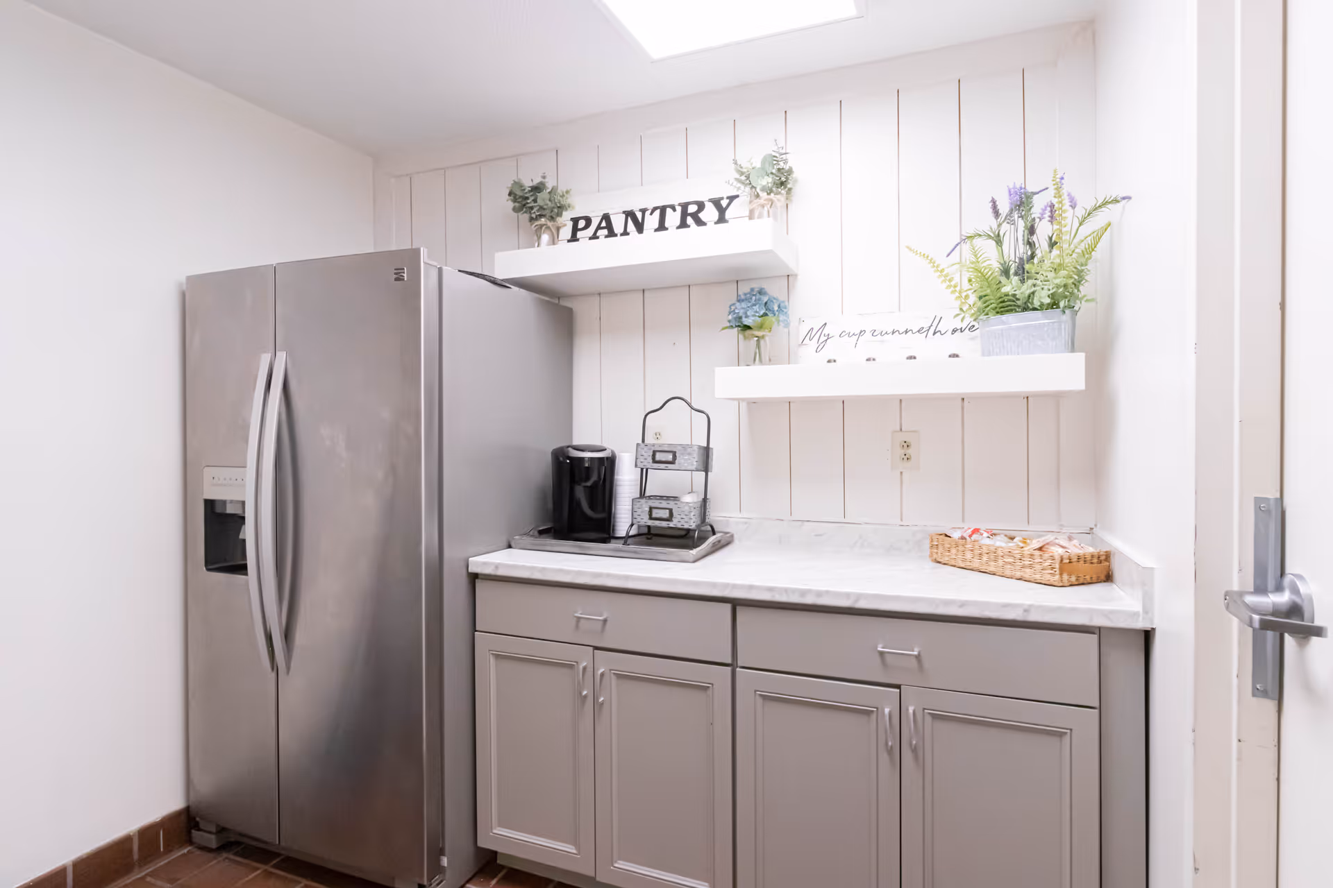 A small pantry area with a stainless steel refrigerator on the left, gray cabinets with a marble countertop, and two white shelves mounted on a white paneled wall. The top shelf has a sign that reads 'PANTRY' and some small potted plants. The lower shelf holds a decorative sign and a larger potted plant. There is a coffee maker and a small basket with packets on the countertop.