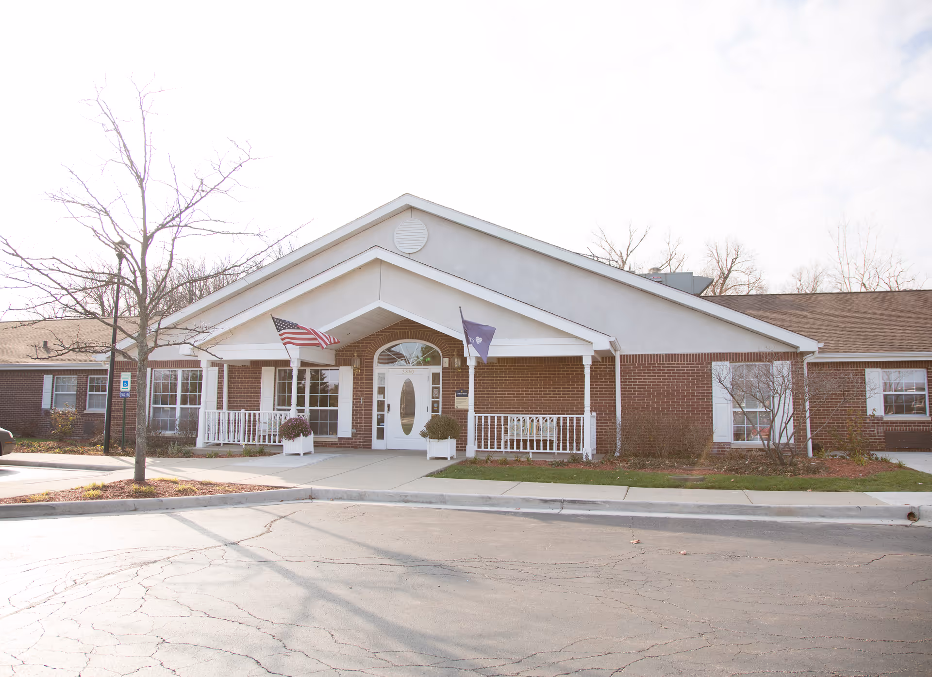 Front exterior view of a single-story brick building with a white entrance porch, two flags on poles near the entrance, and a small tree in front. The building has a paved driveway and sidewalk leading to the entrance.