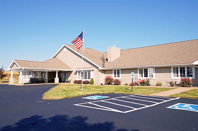 Exterior view of Morningside Assisted Living facility showing a single-story building with beige siding, a covered entrance, an American flag on a flagpole, landscaped bushes and flowers, and two handicap parking spaces in the foreground under a clear blue sky.