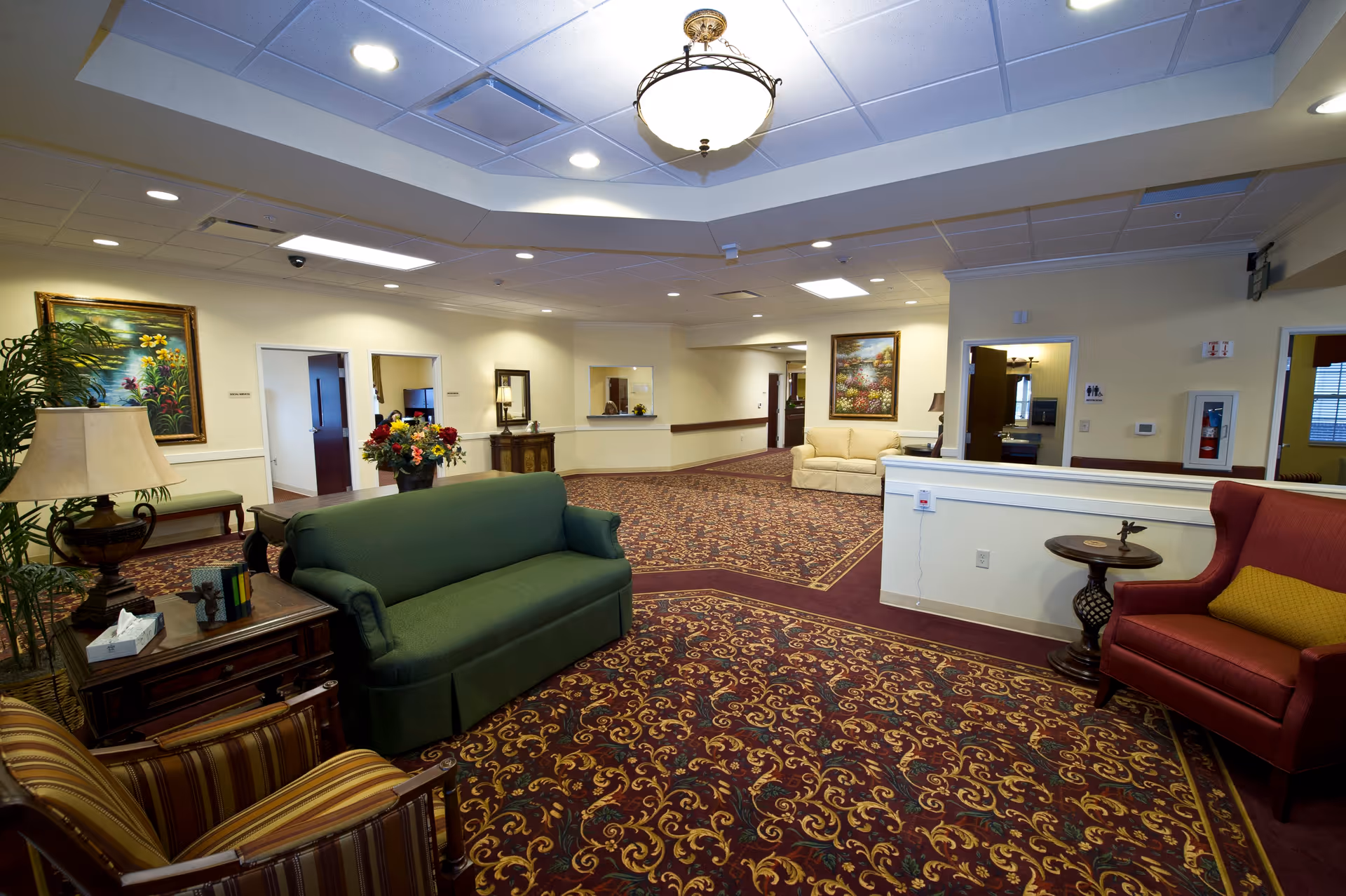 A spacious senior living facility common area with patterned carpet, a green sofa, a red armchair with a yellow cushion, a beige loveseat, and several side tables with lamps and decorative items. The walls are light-colored with framed paintings and there are multiple doorways leading to other rooms. The ceiling has recessed lighting and a central hanging light fixture.