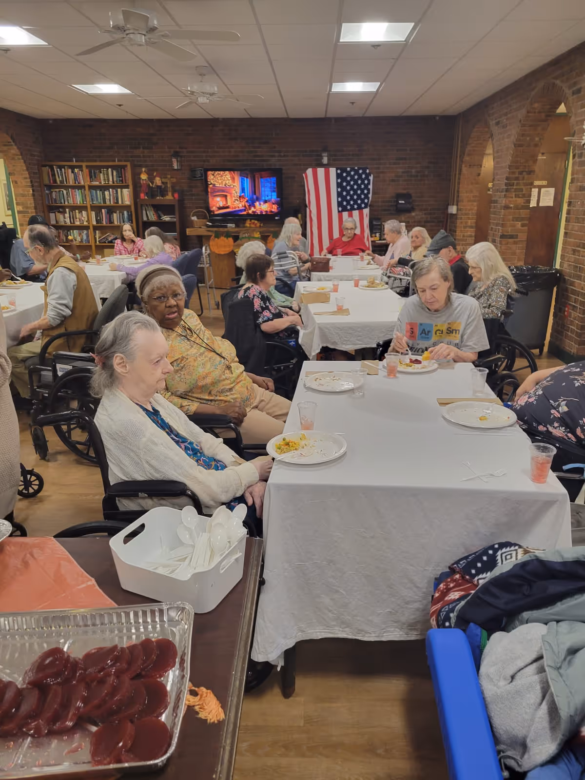 A group of elderly people sitting at tables covered with white tablecloths in a communal dining area. Some are eating while others are conversing. The room has brick walls, a bookshelf, an American flag, and a television displaying a fireplace scene. There are ceiling fans and overhead lights, and a tray of cranberry sauce and utensils is visible in the foreground.