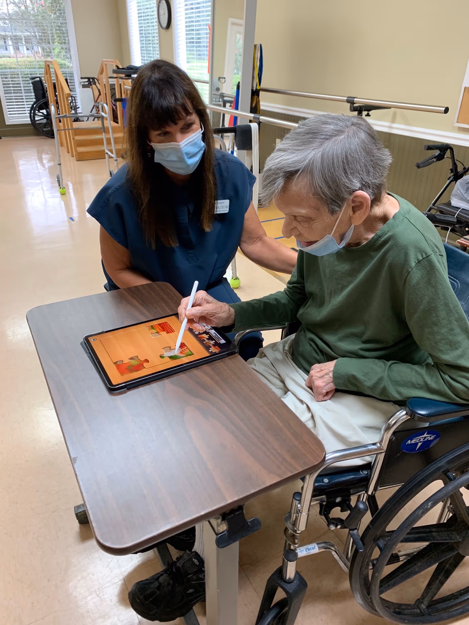 An elderly person in a wheelchair using a stylus to interact with a tablet on a table, while a caregiver wearing a mask watches and supports them in a rehabilitation room with exercise equipment in the background.