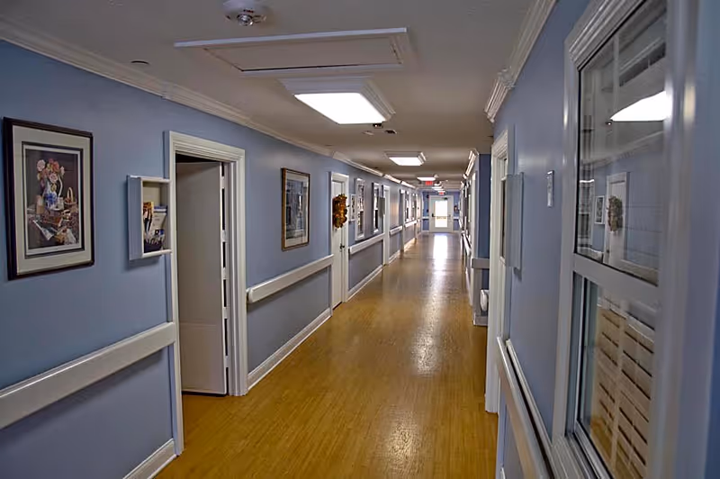 Long indoor hallway with light blue walls and wooden floor, decorated with framed pictures and handrails on both sides, several doors along the corridor, and ceiling lights illuminating the space.