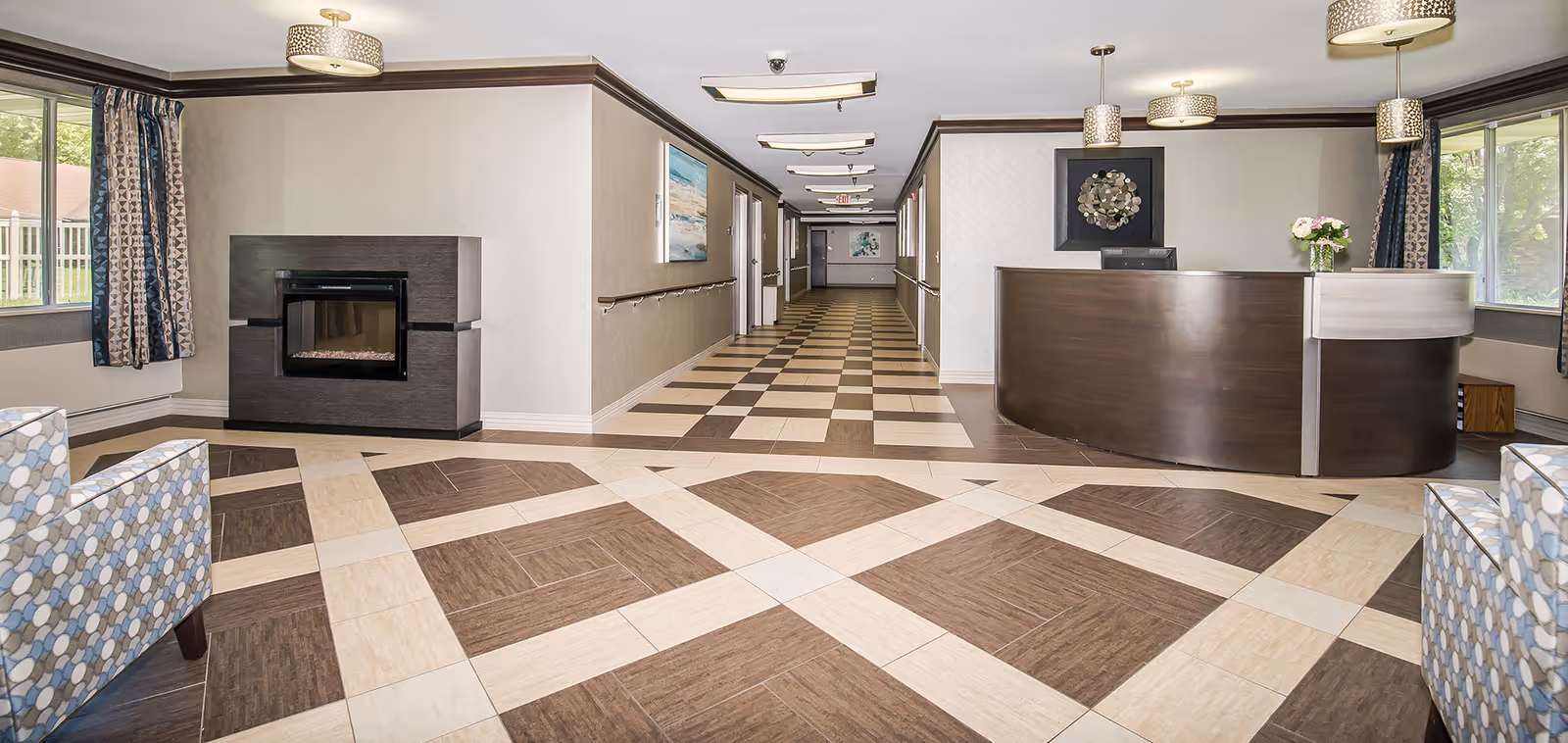 A spacious and well-lit interior hallway of a senior living facility featuring a modern reception desk on the right, a decorative fireplace on the left, patterned tile flooring, and seating areas with patterned armchairs near large windows with curtains.