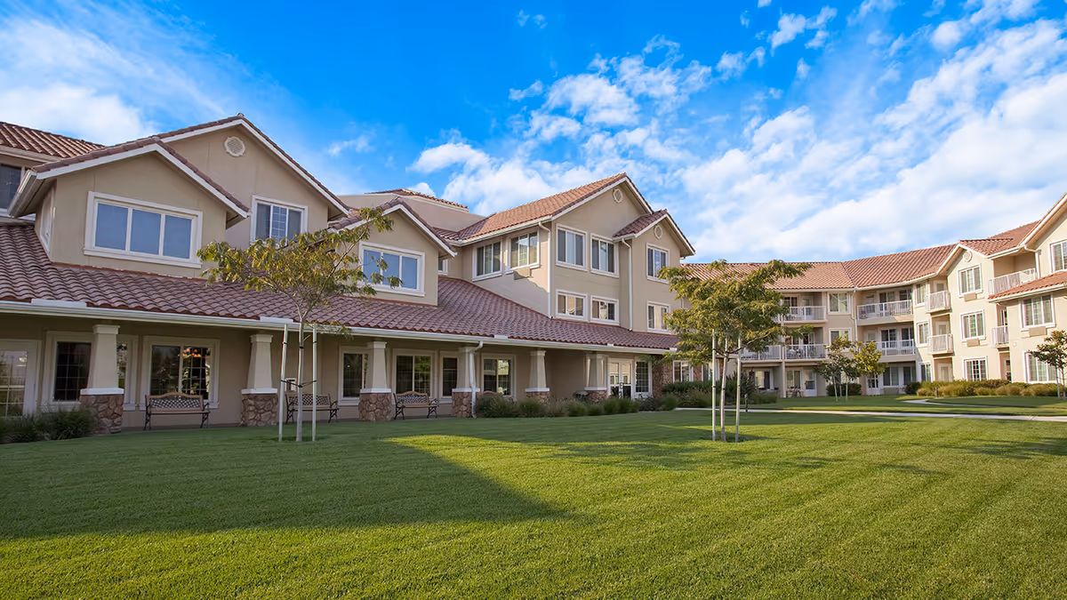 Exterior view of The Orchard Senior Living facility showing a large, well-maintained green lawn with a few small trees and a multi-story beige building with red-tiled roofs and multiple windows under a blue sky with scattered clouds.